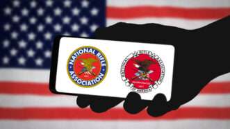 A hand holds a smartphone, displaying two different logos of the National Rifle Assocation (NRA), against a background of the American flag. | Waingro | Dreamstime.com